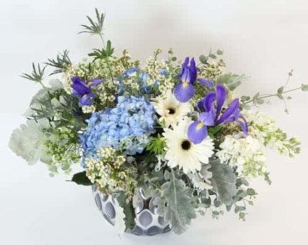 A silver textured bowl holds a gentle design of blue hydrangea, iris, fragrant white stock and white gerbera daisies with accents of waxflower, eucalyptus, dusty miller, eryngium and Queen Anne's lace.