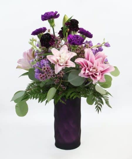This soft romantic design features rose lilies, fragrant stock, purple carnations, scabiosa and accents in a purple vase.