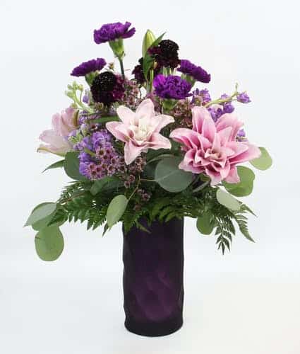 This soft romantic design features rose lilies, fragrant stock, purple carnations, scabiosa and accents in a purple vase. 20"t x 13"w